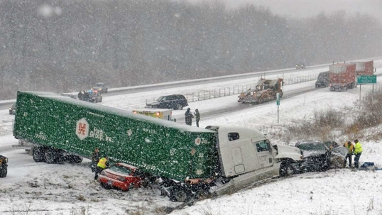 A green semi-truck is crashed into several cars on a snow-covered highway, causing a severe accident.