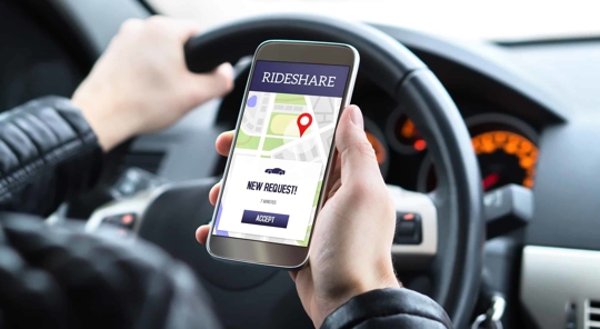 A rideshare driver holds their phone at the steering wheel, with the app displaying a map, pin, car icon, and "New Request" prompt